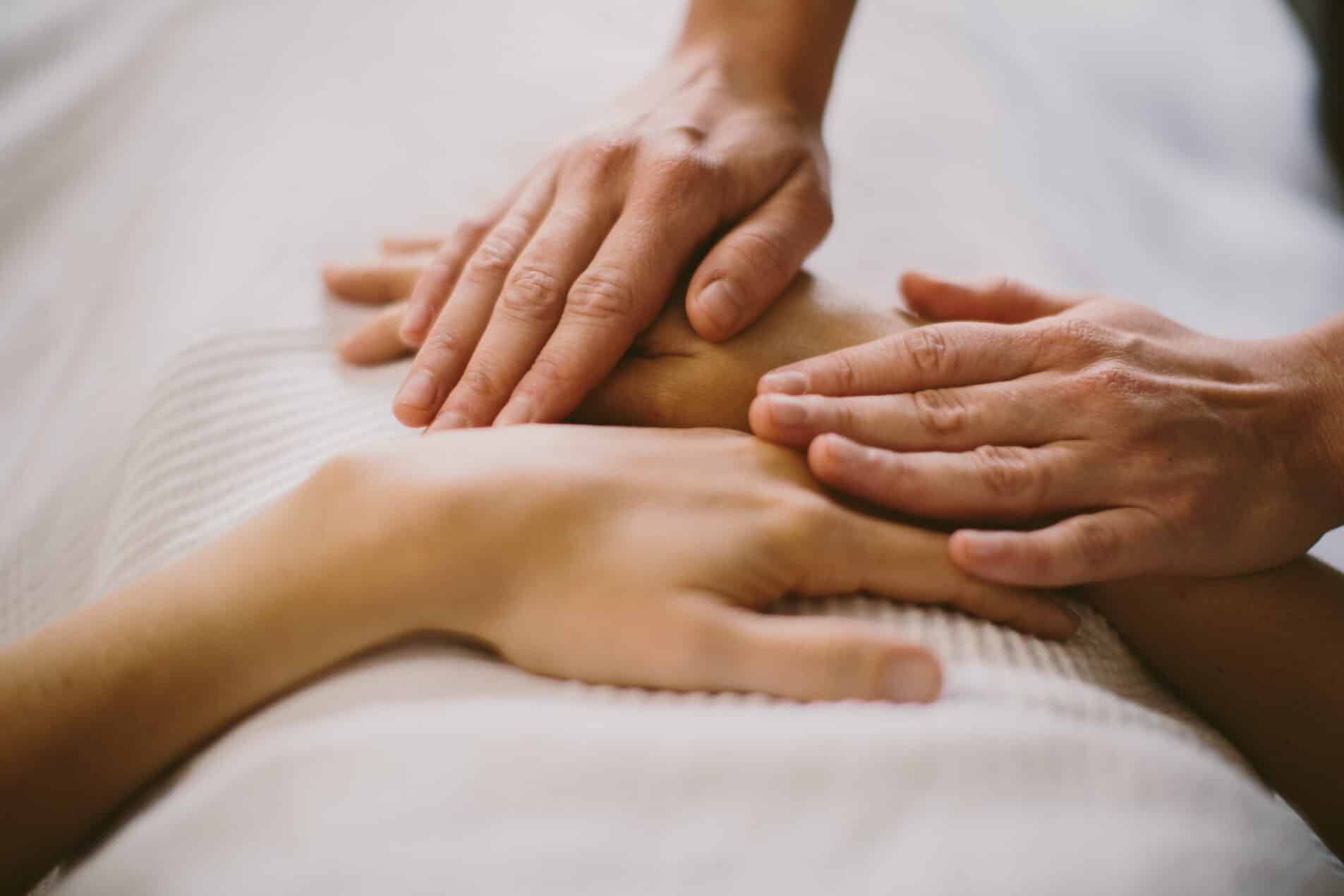 Zwei Paar Hände ruhen auf den Händen und dem Bauch einer Person, die auf einer weißen Unterlage liegt. Die Hände sind sanft übereinander gelegt. Die Szene ist nah und zeigt nur die Hände und einen Teil des Oberkörpers.