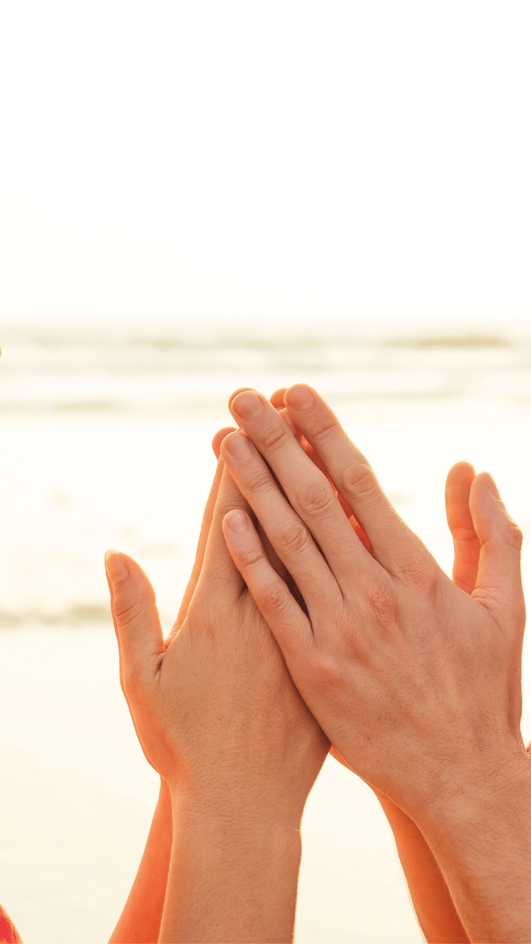 Auf dem Bild sind zwei Hände zu sehen, die an den Handflächen aneinandergelegt sind. Die Hände scheinen von zwei unterschiedlichen Personen zu stammen, und im Hintergrund ist ein heller, unscharfer Strand oder eine Wasserlandschaft sichtbar. Der Fokus liegt auf den Händen, während der Rest des Bildes stark überbelichtet wirkt.