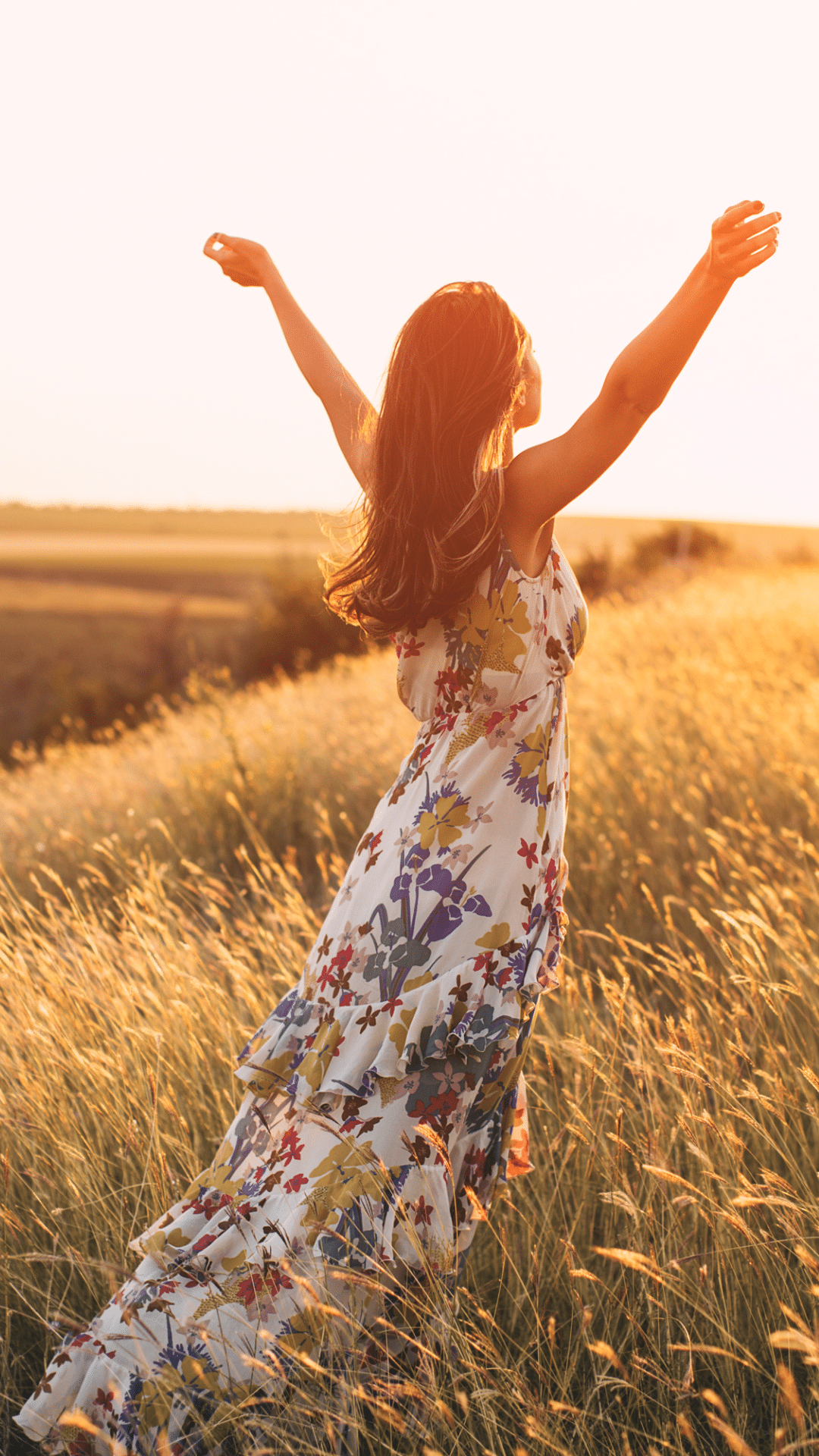 Eine Frau mit langen Haaren steht in einem blühenden Sommerfeld und streckt bei Sonnenuntergang die Arme in die Luft. Sie trägt ein ärmelloses, langes Kleid mit Blumenmuster.