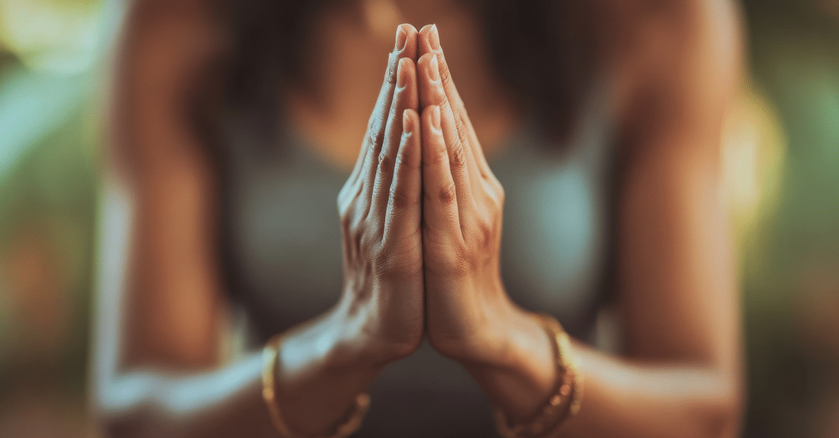Zwei Hände in Gebetshaltung (Anjali Mudra) vor der Brust, scharf fokussiert, mit goldenem Schmuck am Handgelenk. Der Hintergrund ist weichgezeichnet und zeigt eine Person in einem ärmellosen Oberteil in ruhiger Atmosphäre.