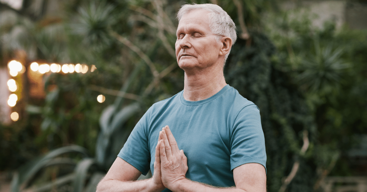 Älterer Mann mit geschlossenem Blick und gefalteten Händen in Gebetshaltung (Anjali Mudra), bekleidet mit einem hellblauen T-Shirt. Er steht inmitten grüner Pflanzen in einem lichtdurchfluteten Raum, vermutlich bei einer Meditations- oder Yogapraxis.