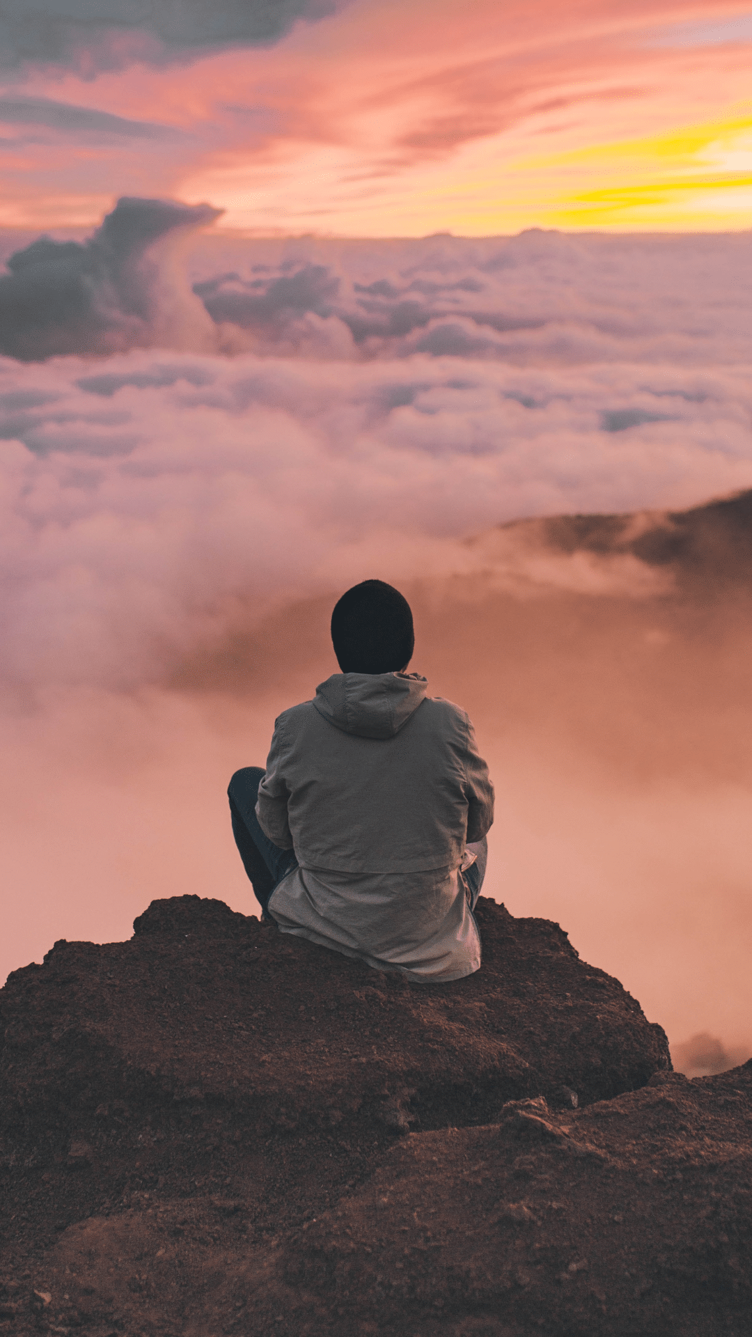 Eine Person sitzt auf einem Felsen und blickt über eine Wolkendecke bei Sonnenuntergang.