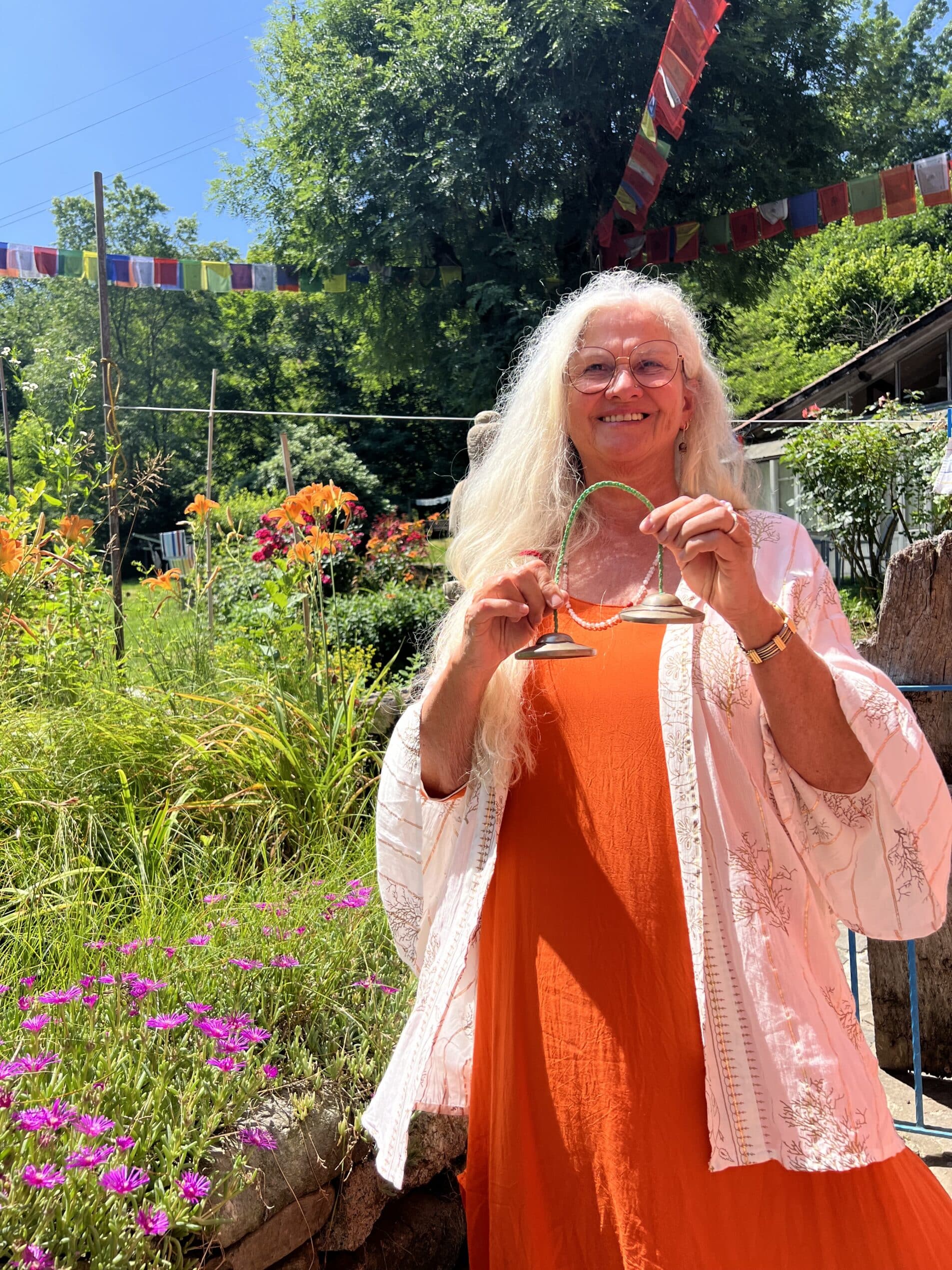 Das Bild zeigt Moti Theresia König stehend während eines Seminars in Molino. Hinter ihr ist steht ein Baum und um sie herum ist viel grünes Gras und sie trägt ein orangenes Kleid.
